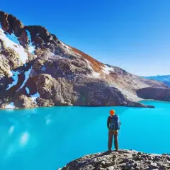 Sender solitario contempla un lago turquesa en la patagonia chilena, rodeado de montañas nevadas y un cielo azul intenso.
