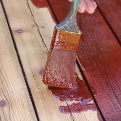 Application d'une teinture rouge sur des planches de bois. Un pinceau chargé de produit montre comment peindre du bois pour le protéger.