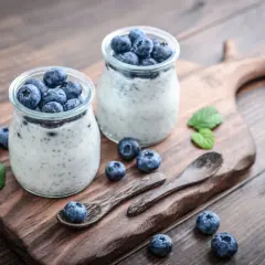 Dwa słoiczki z deserem pudding chia, ozdobione świeżymi borówkami i listkami mięty, na drewnianej desce.