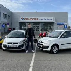 Un jeune homme pose devant une auto-école avec deux voitures sans permis et des scooters. Il tient deux casques, prêt pour son apprentissage.
