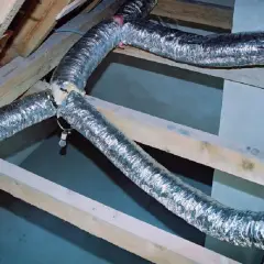 Elastyczna, aluminiowa rura doprowadzająca powietrze do kominka, zamontowana pod dachem na drewnianych belkach.
