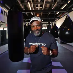 A man in a "UNDEFATED" shirt and Mayweather Boxing + Fitness hat poses with fists up in a gym, ready for Floyd Mayweather training.