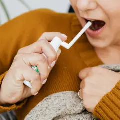 Une personne utilise un spray buccal pour soulager un mal de gorge. Elle tient le flacon blanc et dirige l'embout vers sa bouche ouverte.