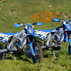 Zwei blaue Enduro-Motorräder, ein 2-Takter und ein 4-Takter, stehen auf einer grünen Wiese.