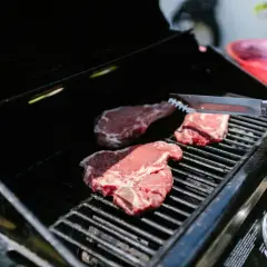 Trzy soczyste steki na ruszcie grilla, gotowe do grillowania. Dłoń z szczypcami zbliża się do mięsa.
