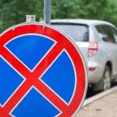 Znak zakazu zatrzymywania się b36. Samochody stoją w rzędzie przy ulicy, ale ten znak przypomina, że nie wolno tu parkować.