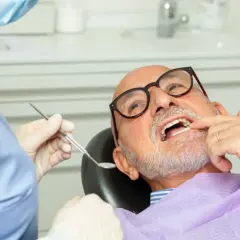 Dentysta w masce i rękawiczkach bada uśmiech starszego mężczyzny w okularach.