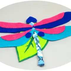 Libélula de fieltro, un proyecto de manualidades con alas azules, rosas y turquesas, posada sobre una hoja verde.