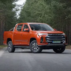 Pomarańczowy Maxus T90 na leśnej drodze. Ten potężny pickup z charakterystycznym grillem i nowoczesnymi światłami świetnie prezentuje się w otoczeniu drzew.