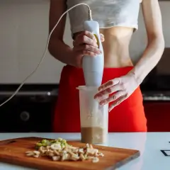 Kobieta miksuje składniki na shake'a blenderem ręcznym. Na desce pokrojone owoce.