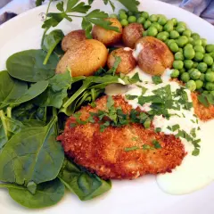En panerad schnitzel med krämig sås, serverad med färsk spenat, små potatisar och gröna ärtor. En klassisk rätt där såsen till schnitzel lyfter hela smakupplevelsen.