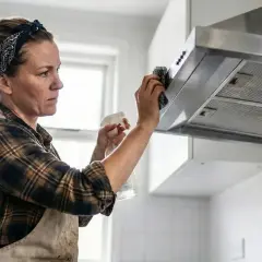 Kobieta w fartuchu i chustce na głowie czyści okap kuchenny. Pokazuje, jak wyczyścić okap z tłuszczu, używając sprayu i gąbki.