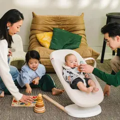 Familjen leker på golvet. Bebisen sitter i en Stokke babysitter medan pappan leker med den. Systern leker med en träleksak.