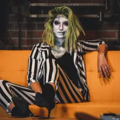 Donna con trucco spettrale e capelli verdi, indossa un vestito da strega fai da te a righe bianche e nere, seduta su un divano arancione.