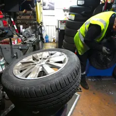 Mechanik w kamizelce odblaskowej pracuje przy kole. Słychać buczenie opon podczas jazdy, gdy pracownik wymienia oponę.