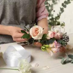 Osoba w fartuchu tworzy bukiet z różowych i kremowych kwiatów, obok leży biała wstążka.