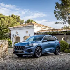 Niebieski Ford SUV Kuga, jeden z popularnych ford suv modele, zaparkowany na brukowanym podjeździe przed białym domem z czerwoną dachówką.