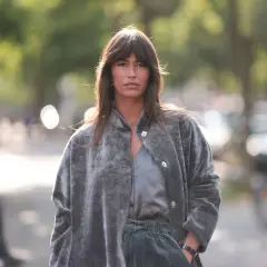 Frau mit curtain bangs und natürlichen Locken trägt einen grauen Kunstpelzmantel und grüne Hose.