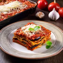 Una fetta di lasagne vegetariane, con melanzane e pomodoro, guarnita con basilico e parmigiano. Sullo sfondo, ingredienti freschi.