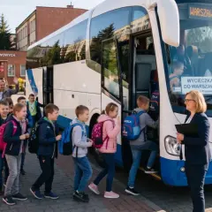 Dzieci z plecakami wsiadają do autobusu na wycieczkę do Warszawy. W tle szkoła i rodzice machający na pożegnanie.