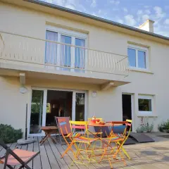 Maison divisée en 2 logements avec terrasse en bois, mobilier coloré et transat. Jardin verdoyant et ciel bleu.