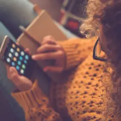 Kobieta w okularach i swetrze trzyma telefon z ikonami aplikacji i notatnik.