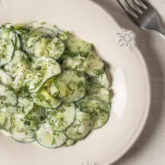 Ensalada alemana de pepino en rodajas finas, cubierta con una cremosa salsa y eneldo fresco, servida en un plato blanco.