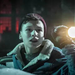 Chłopiec w zimowej kurtce i czapce trzymający latarkę, oświetla mroczny, ośnieżony teren. Może szuka czegoś w pobliżu czarnego młyna.