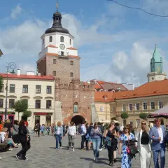 Ludzie spacerują po brukowanym placu litewskim, podziwiając zabytkową wieżę z zegarem i zieloną iglicę w tle.
