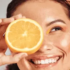 Strahlendes Lächeln mit einer Orangenscheibe vor dem Auge, die Frische und Vitamin C für die Hautpflege symbolisiert.
