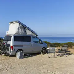 Silberner Camper mit Dachzelt und Fahrrädern am Heck, zwei Liegestühle und ein Tisch stehen bereit für entspanntes italien wildcampen am Meer.