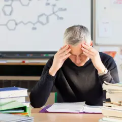 Lehrer mit Händen an den Schläfen, umgeben von Büchern und einer chemischen Formel an der Tafel. Der Lehrer-Stress ist deutlich sichtbar.