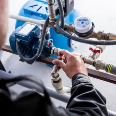 Ręka majstra reguluje zawór na grupie pompowej co. W tle niebieski zbiornik i wodomierz.