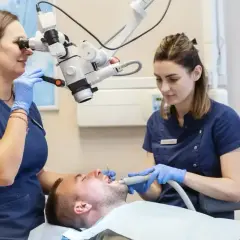 Dwie dentystki w gabinecie stomatologicznym pracują przy pacjencie, jedna używa mikroskopu, druga narzędzia.