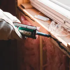 Hand med handske applicerar tätningsmassa för att ta bort silikonfog runt ett fönster.