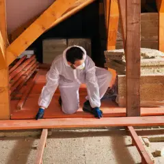 Un ouvrier en combinaison blanche installe des planches de bois pour refaire un plancher sur poutre, entouré de matériaux d'isolation.