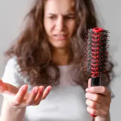 Mujer preocupada muestra mechones de pelo en su mano y un cepillo con más cabello, buscando un tratamiento para pelo que se rompe.