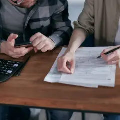 Dwie osoby przy drewnianym stole wypełniają dokumenty, obok kalkulator i kubek.