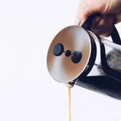 Dłoń trzyma zaparzacz french press, z którego właśnie leje się aromatyczna kawa.