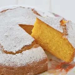 Una fetta di torta di carote e yogurt, soffice e dorata, viene sollevata da una spatola. La torta è cosparsa di zucchero a velo.
