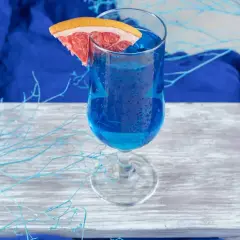 Niebieski drink, jak orzeźwiająca blue lagoon, z plasterkiem grejpfruta, na drewnianej desce, otoczony błękitnymi gałązkami.