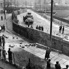 Żołnierze pilnują muru, który powstał, by podzielić Berlin. Ludzie obserwują z dystansem.