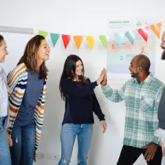 Ein motiviertes Lehrerteam feiert einen Erfolg mit High-Fives. Bunte Wimpelketten und eine Wandtafel deuten auf eine positive Lernumgebung hin.