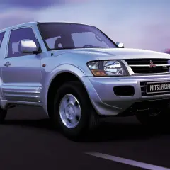 Srebrny Mitsubishi Shogun 3-drzwiowy na drodze o zmierzchu, z miejskim krajobrazem w tle.
