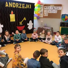 Dzieci na andrzejkach w przedszkolu, siedzą w kółku. W tle dekoracje z napisem "ANDRZEJKI", świece i duchy.