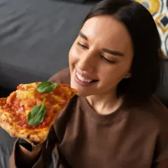 Uśmiechnięta kobieta delektuje się kawałkiem pizzy, jakby właśnie zakończyła jej odgrzewanie w piekarniku.
