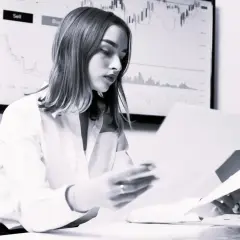 Kobieta analizuje dokumenty, podczas gdy na ekranie w tle widać wykresy finansowe. Bank sprawdza co po udzieleniu kredytu, by upewnić się co do stabilności finansowej.