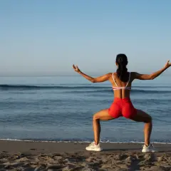 Kobieta w czerwonych szortach i sportowym topie ćwiczy na plaży. Jej szeroko rozstawione nogi i wyciągnięte ręce sugerują rozciąganie, być może angażujące powięź szeroką.