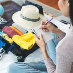 Kobieta pisze listę rzeczy, co zabrać na wakacje all inclusive: paszport, aparat, kapelusz, ubrania, poduszkę podróżną.