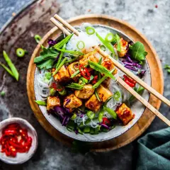 En skål med friterade glasnudlar, tofu, rödkål och gröna lökar, toppad med chili och serverad med ätpinnar.